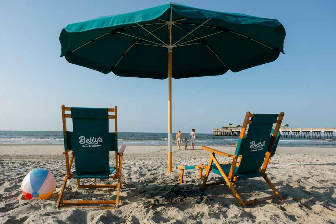 Two teal beach chairs with wooden frames under a large teal umbrella on a sandy beach. The left chair has a colorful beach ball next to it, and both chairs display the brand name 'Betty's'.